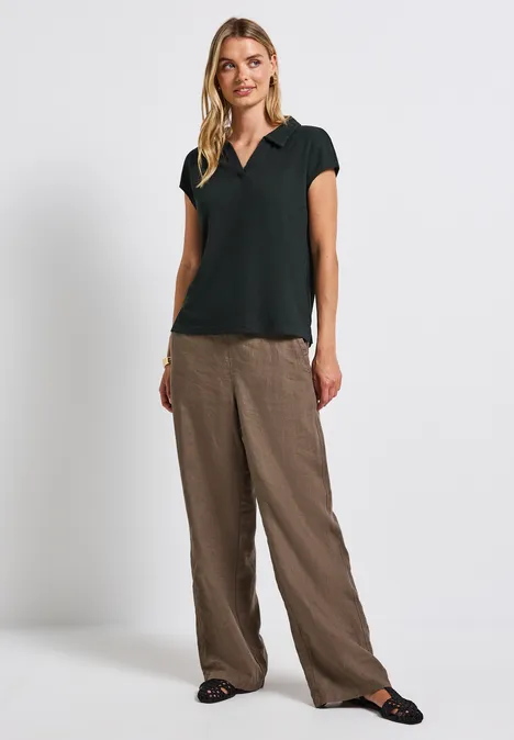 Frau mit schwarzem Shirt und brauner weiter Hose steht vor weißem Hintergrund.